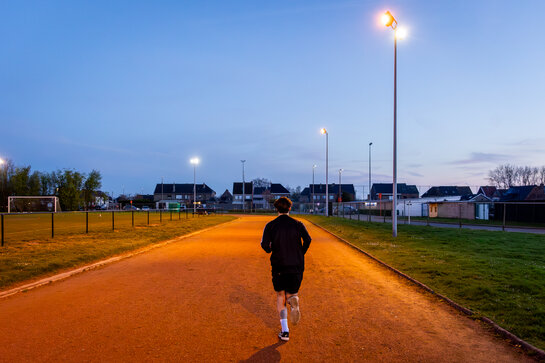 Jogger op een goed verlichte looppiste naast een sportterrein tijdens de schemering.