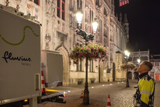 Straatverlichting in een stadskern, met Fluvius-voertuig en verlichtingspalen voor een monumentaal gebouw.