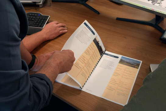 Wil je ook een masterplan openbare verlichting opstellen? Persoon bladert door een document met schema’s en richtlijnen voor een masterplan openbare verlichting op een bureau met computerschermen en werkmateriaal.